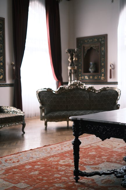 A vintage-style living room featuring ornate wooden furniture with intricate carvings, including a dark upholstered sofa and a matching armchair. The room has large windows with sheer white curtains a